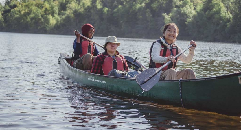 three people wearing life jackets paddle a canoe toward the camera. The person in the front of a canoe smiles at the camera. 
